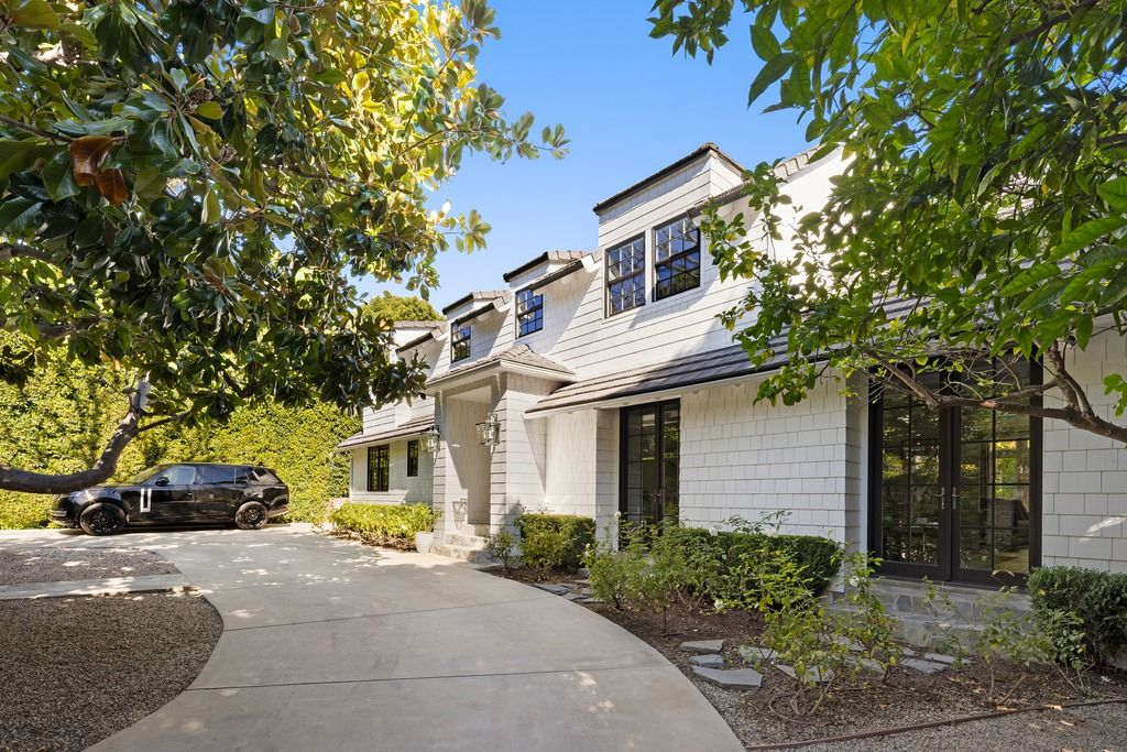 White shingle-style siding, black-framed windows, and a series of dormers give the exterior a clean East Coast-inspired look, while tall hedges and magnolia trees soften the entrance and add privacy from the street.