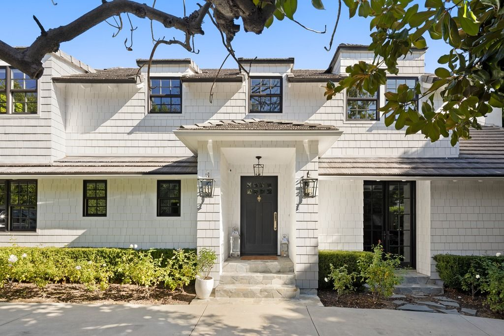 As the driveway widens near the entrance, a covered front porch sits beneath the upper level, framed by lantern-style lighting and dark double doors that contrast against the lighter exterior palette.