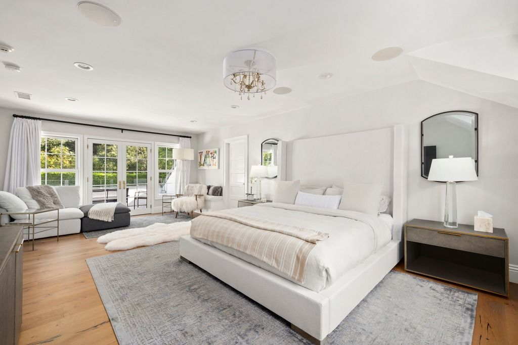 The bedroom centers around a tall upholstered bed framed by soft gray walls, warm wood flooring, and oversized rugs that help define the seating and sleeping areas.