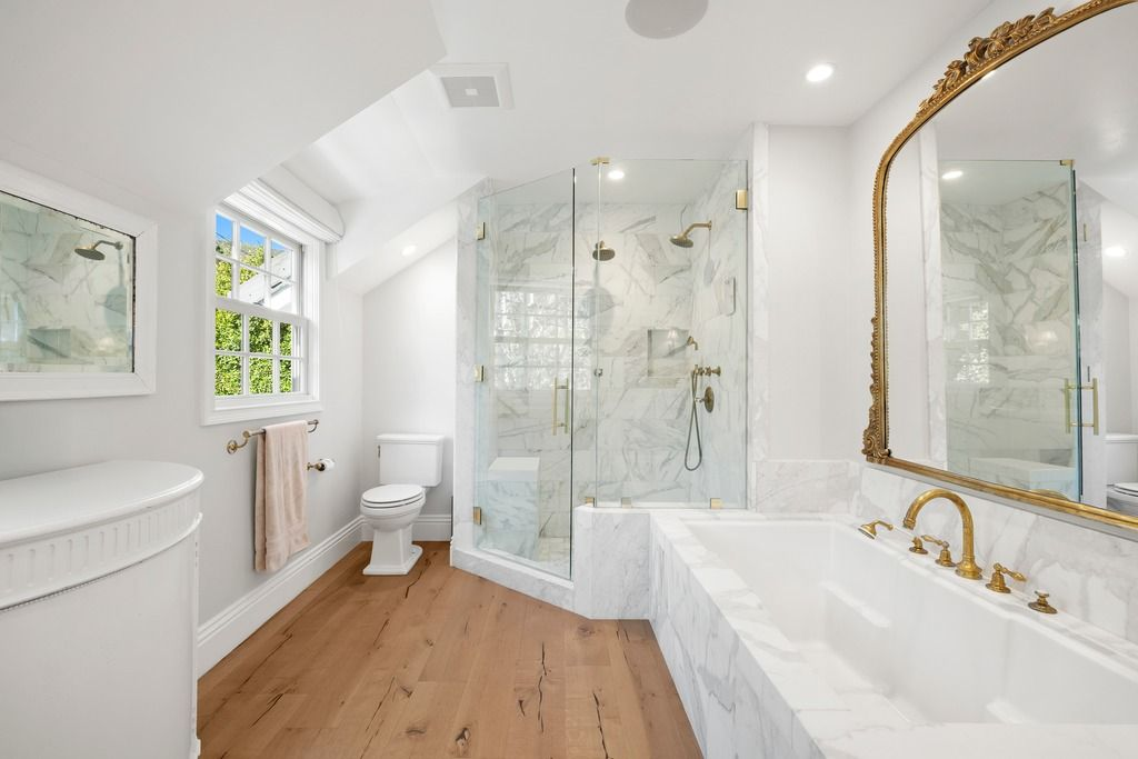 A soaking tub sits beneath an oversized mirror, while the shower incorporates marble slab walls, built-in shelving, and updated hardware throughout.