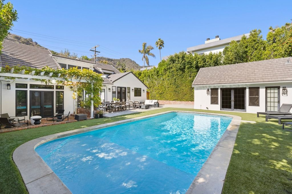 The pool anchors the center of the backyard, bordered by grass lawns, lounge chairs, and a detached guesthouse.