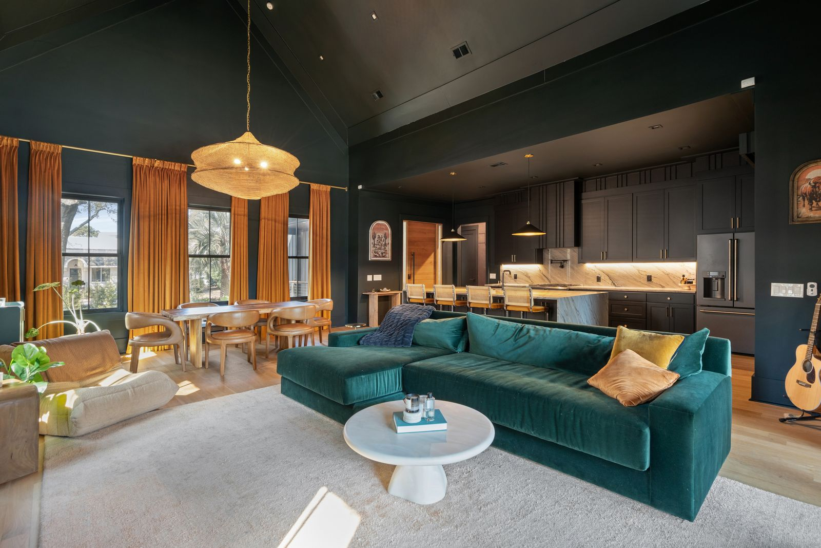Inside, the home opens into a double-height living room beneath soaring vaulted ceilings and matte black walls, while oversized rust-colored drapes stretch upward beside tall windows, emphasizing the room’s vertical scale and drawing natural light into the richly layered design.