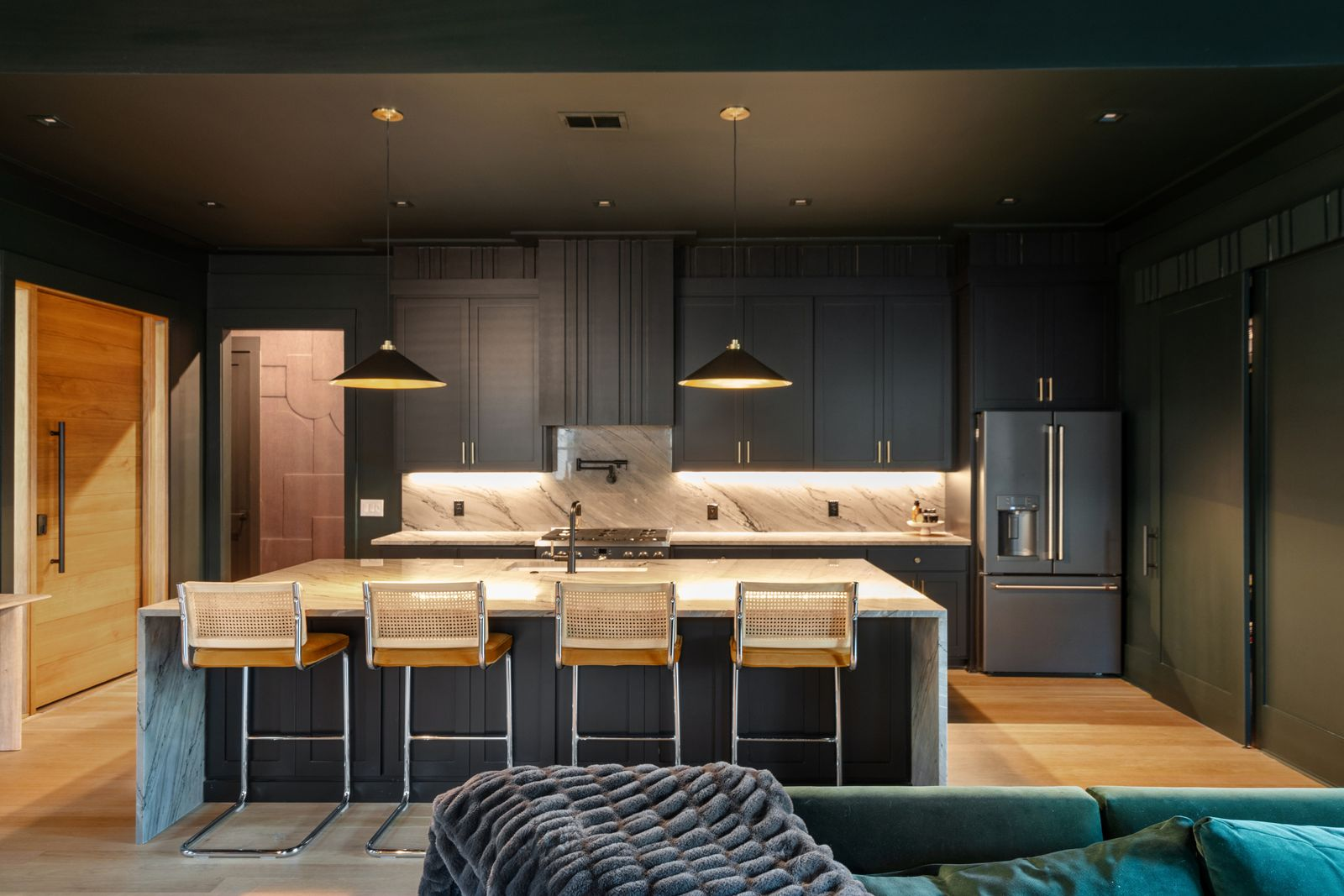 The space is centered around a massive waterfall island wrapped in heavily veined stone, creating a sharp contrast against the darker finishes with woven bar seating tucked beneath the counter.