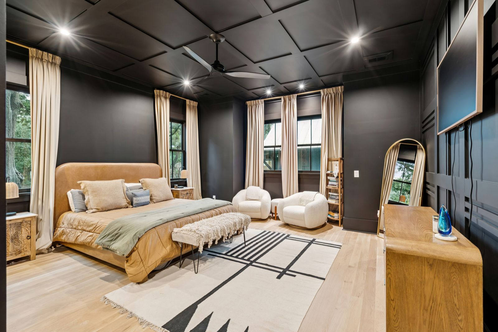 Beginning the suite is the bedroom, which centers around a tall upholstered bed framed by matte black walls, warm wood flooring, and layered neutral fabrics. 
