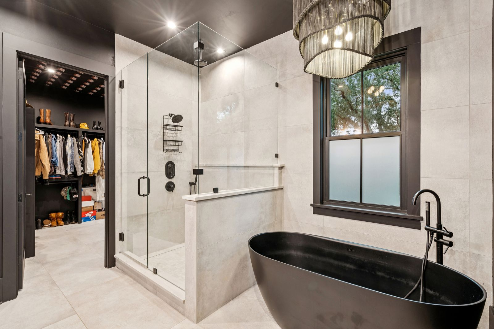 Across from the vanity, a glass-enclosed walk-in shower sits next to a freestanding black soaking tub positioned beneath a window overlooking the surrounding trees.