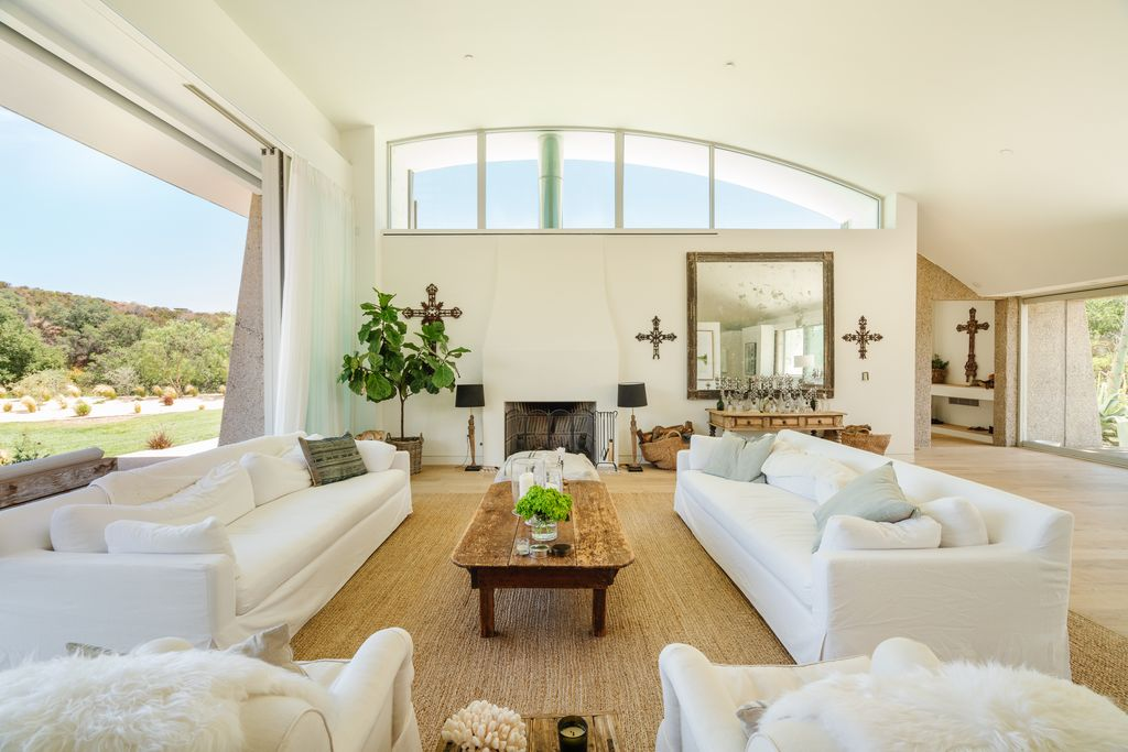 The room is arranged around a central fireplace framed by a tall white chimney and oversized clerestory windows, while wide sliding glass openings stretch across the space.