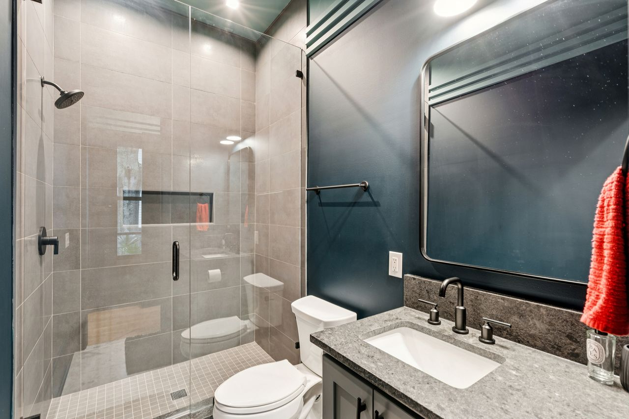 Its compact en-suite bathroom continues the suite's deep blue walls, featuring matte black fixtures, a stone-topped vanity, and large-format gray tile inside the glass-enclosed shower.