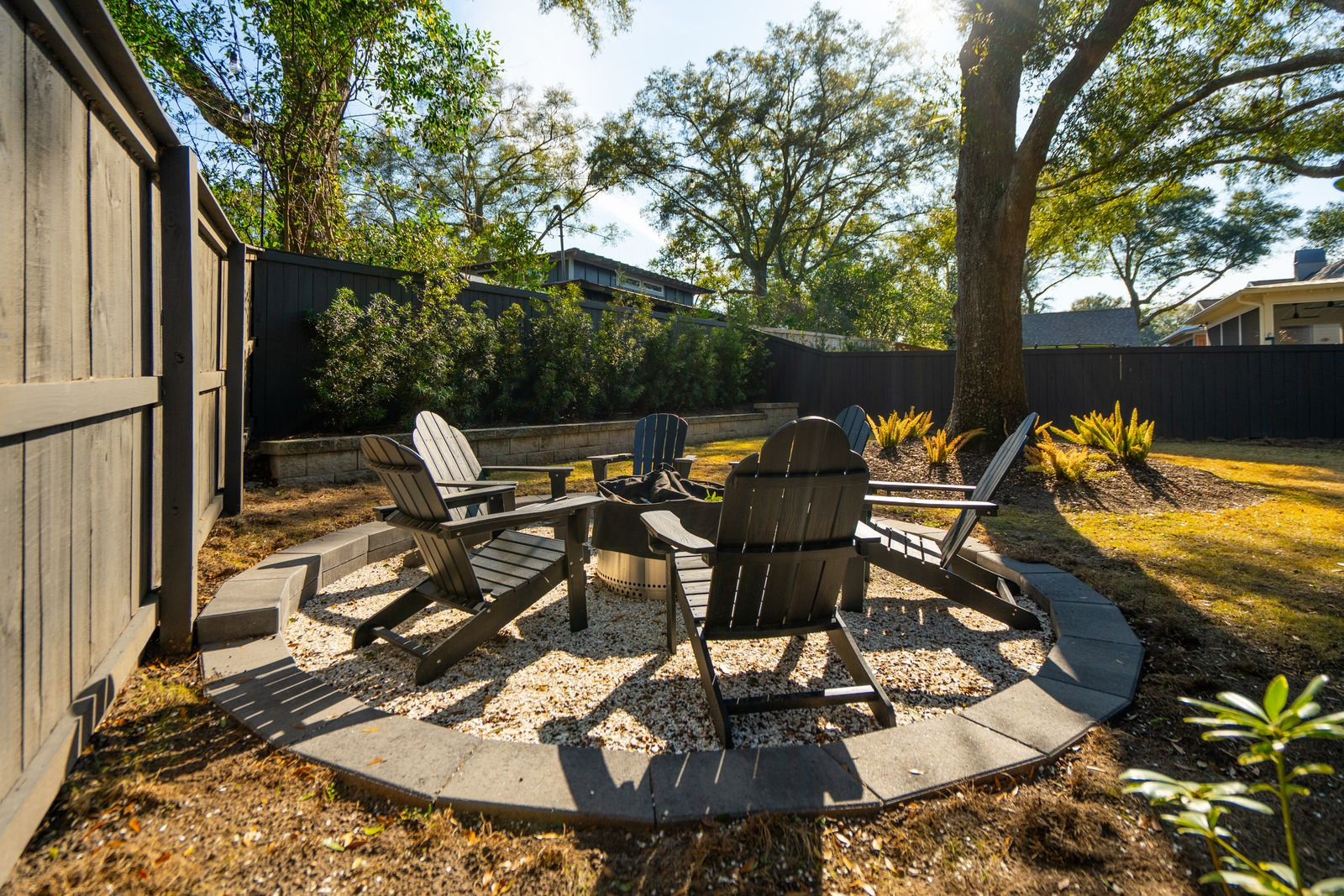 Off to one side of the yard, a circular gravel firepit lounge creates a gathering space beneath the trees, surrounded by Adirondack chairs.