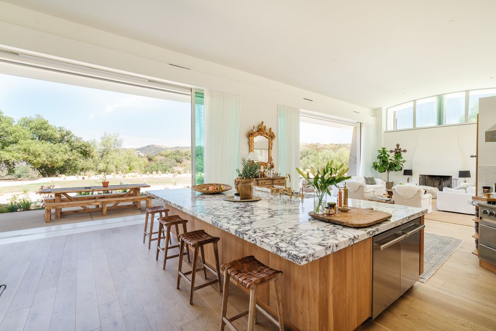 The kitchen is framed by soaring ceilings and oversized glass openings that slide away to connect the interior directly to the outdoor dining terrace and canyon backdrop beyond.