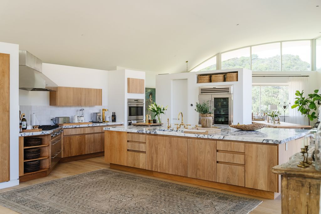A massive marble-topped island anchors the space, stretching nearly the length of the room with integrated seating, prep areas, and brass fixtures that add warmth against the natural oak cabinetry.