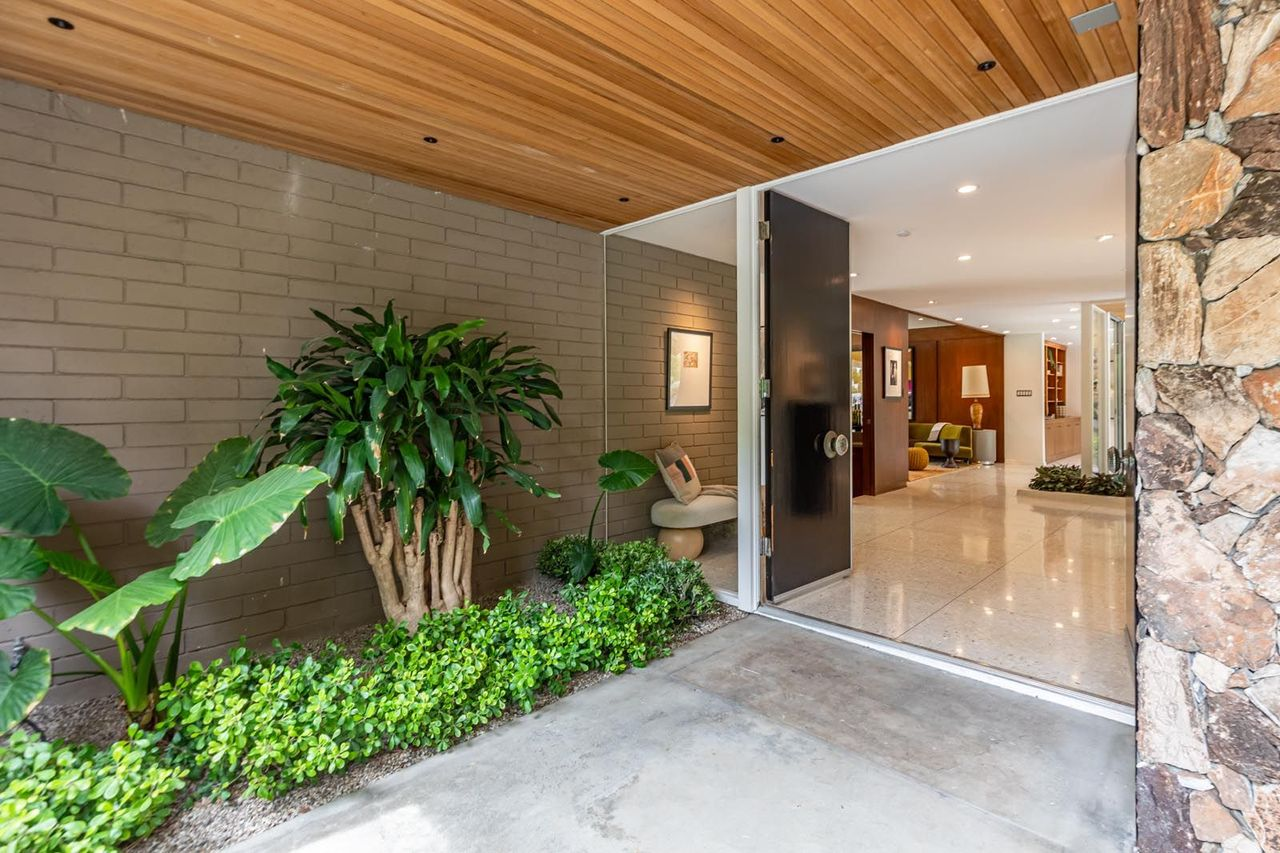 Entering the home, a covered entryway framed by walls of grey brick and natural stone guides visitors past built-in planter beds and toward the oversized entry doors.