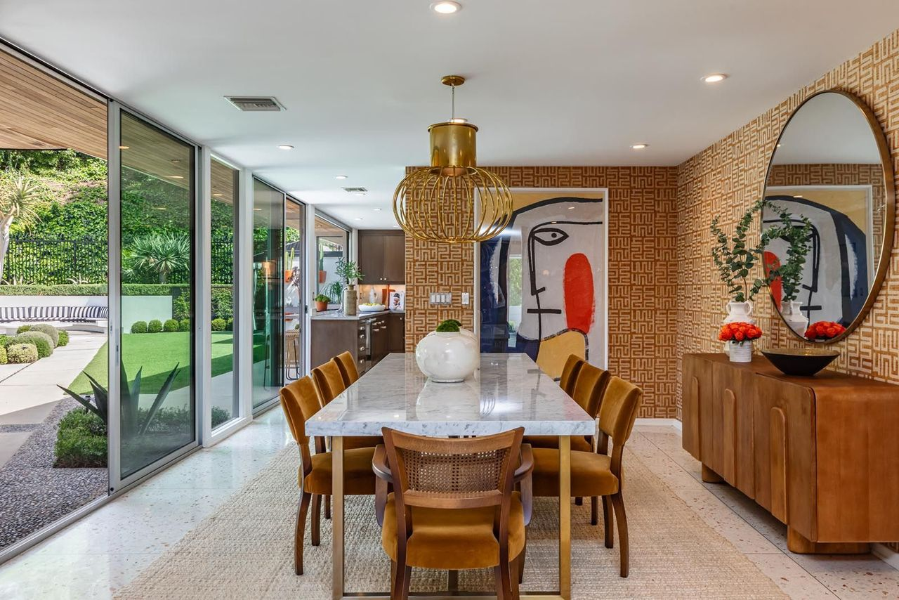Around the corner, the dining room continues the home’s open mid-century layout with a long marble table surrounded by velvet upholstered chairs beneath a brass statement chandelier.