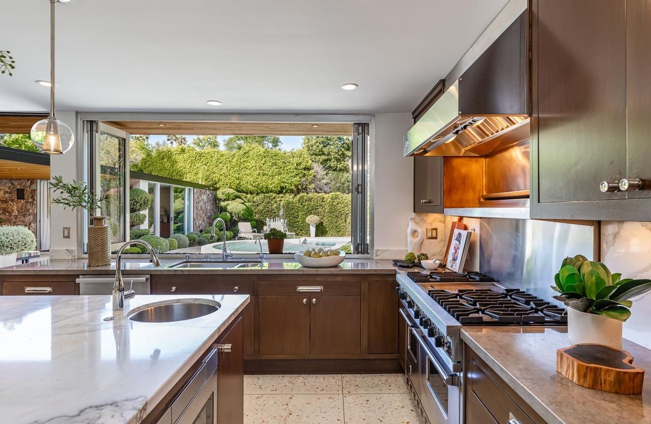 Professional-grade stainless steel appliances are integrated into the cabinetry wall, including a large commercial-style range and vent hood, while long counters wrap around the perimeter beneath expansive windows.
