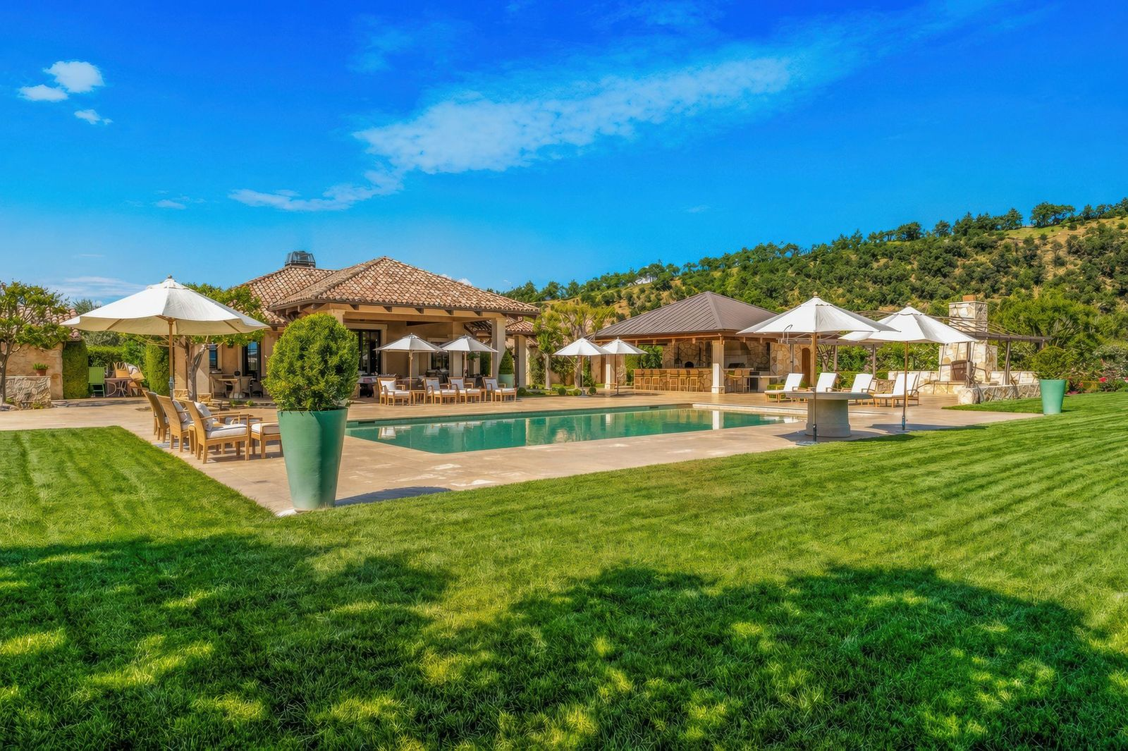 Positioned beside the main residence and framed by rolling hills in the distance, the setup has the scale and layout of a private resort, with multiple shaded seating areas spread throughout the deck.