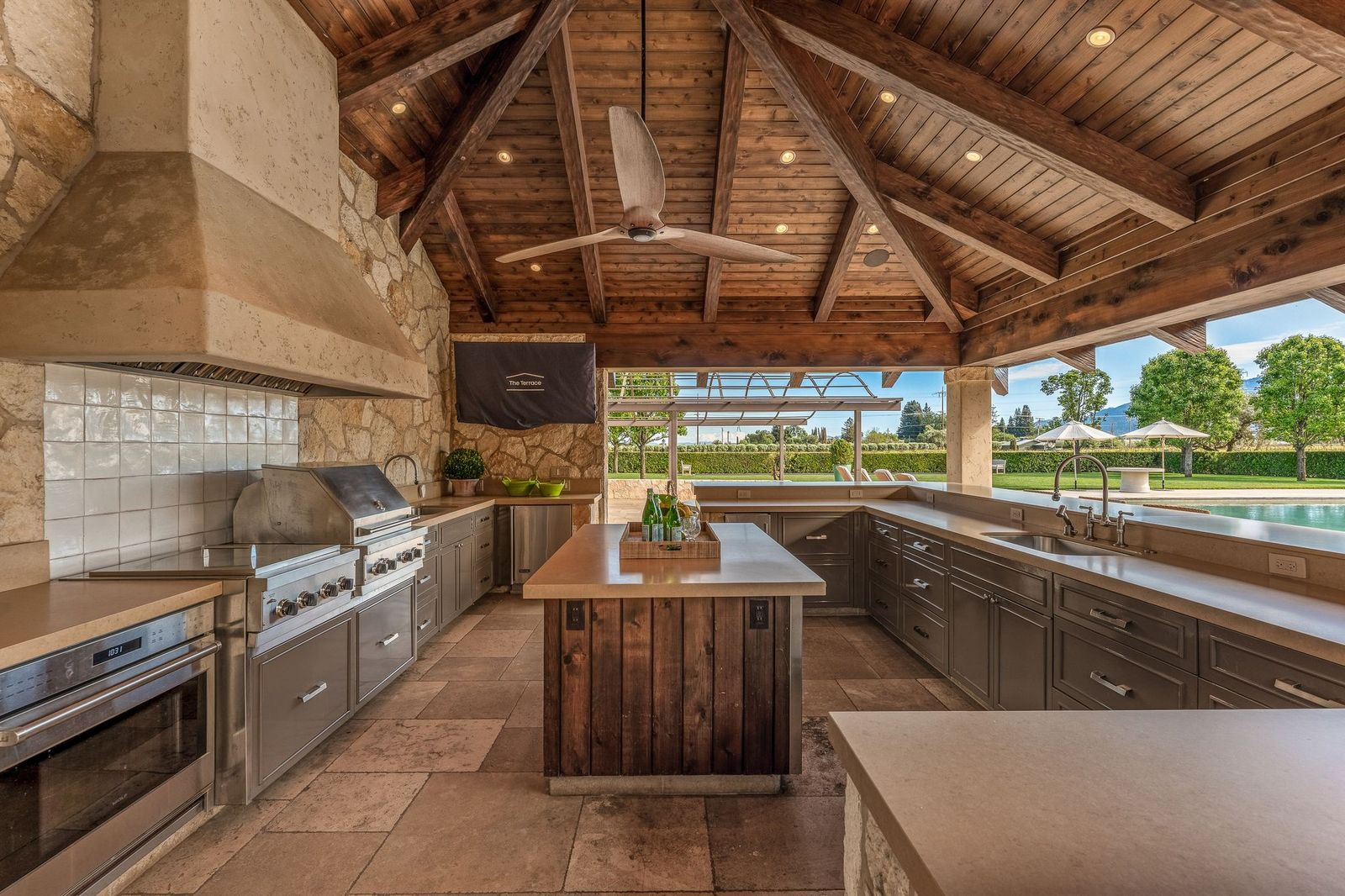 A few steps away, the outdoor kitchen pavilion was built for hosting, featuring a full grilling station, oversized prep counters, integrated appliances, and a central island beneath exposed timber trusses.