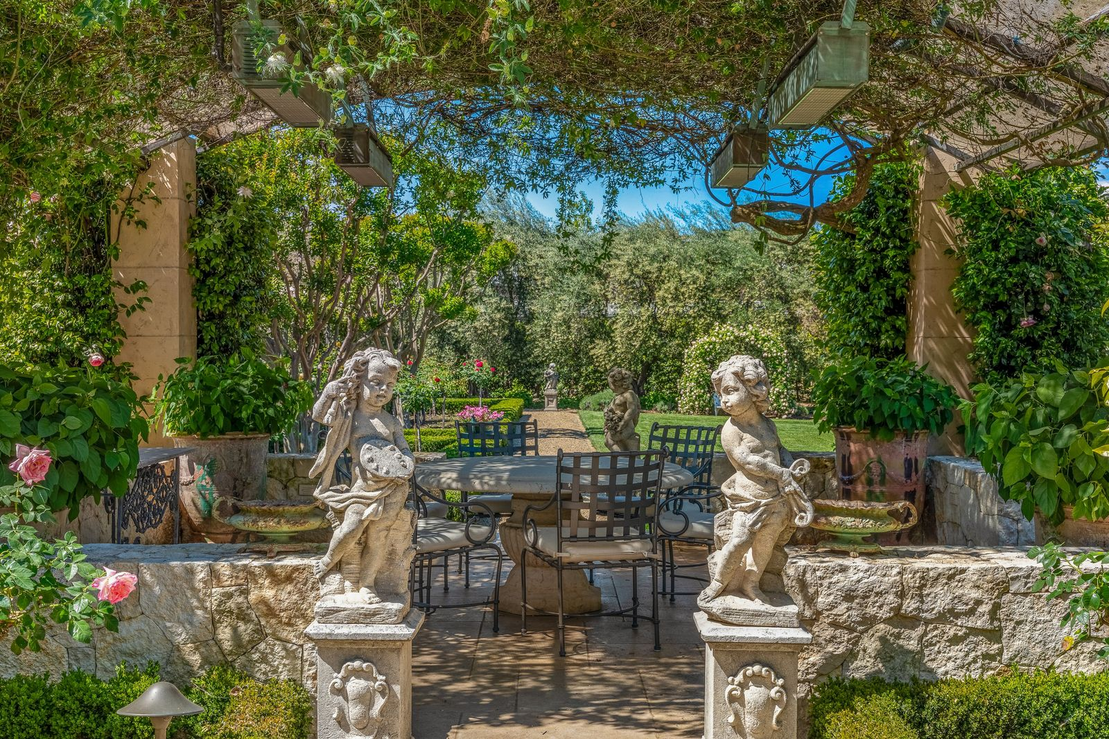 Several garden pathways and pergola-covered seating spaces are woven throughout the grounds, including formal dining terraces framed by stonework, sculptures, and climbing vines overhead.