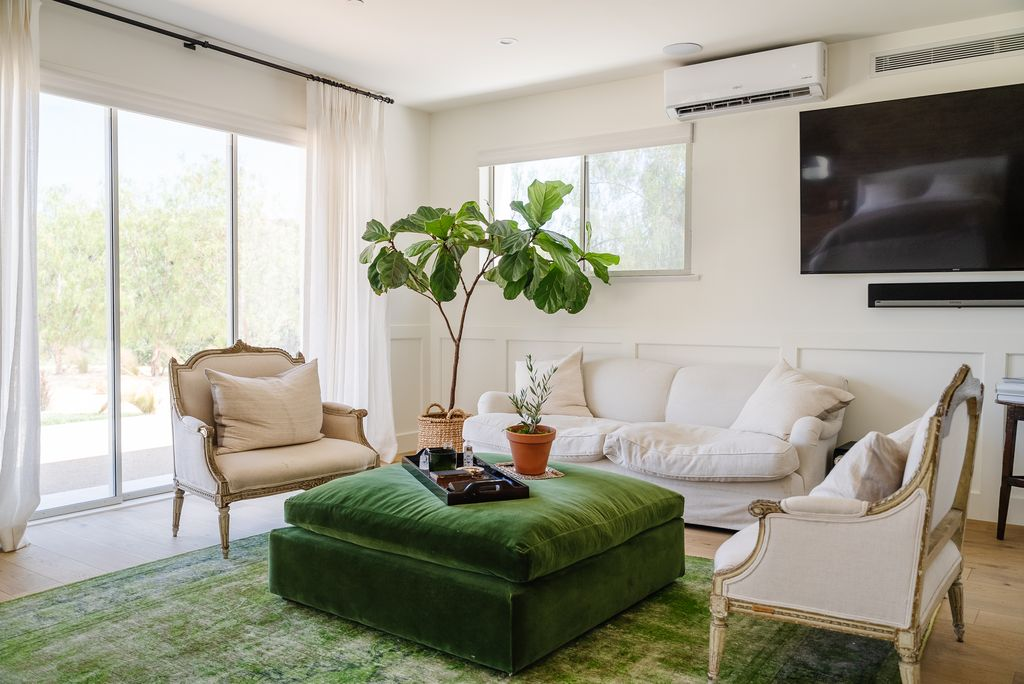 Across from the bed, the lounge area consists of a low white sofa and vintage-style armchairs arranged around a large green ottoman.
