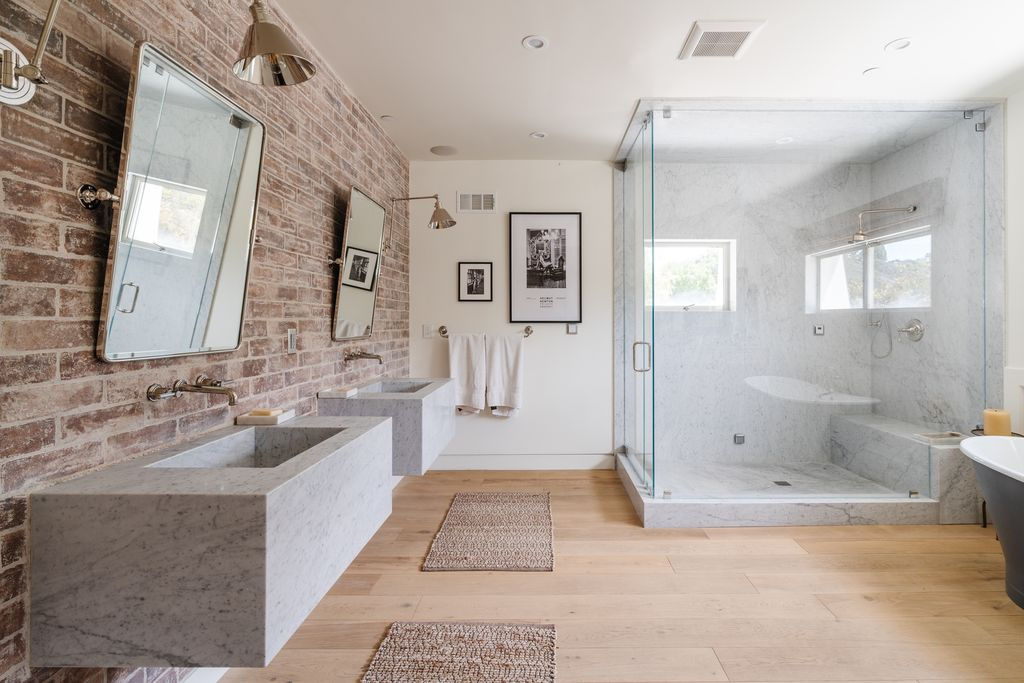 A large glass-enclosed shower occupies one side of the room, wrapped almost entirely in marble, while floating stone sinks line the opposite brick wall beneath industrial-style mirrors and sconces.