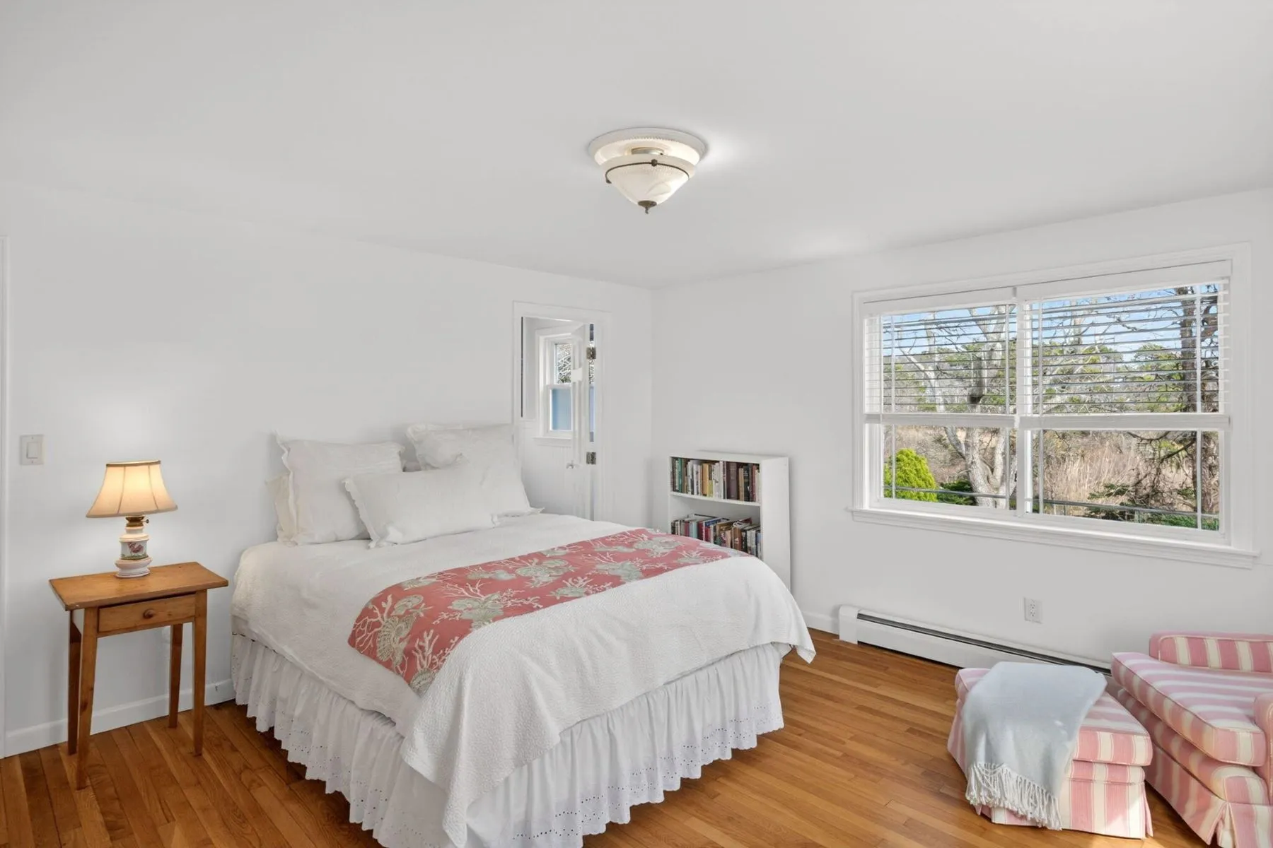 One of the bedrooms features a clean, airy setup centered around a white bed layered with soft linens and a coral-patterned throw that adds a coastal touch to the neutral space. 