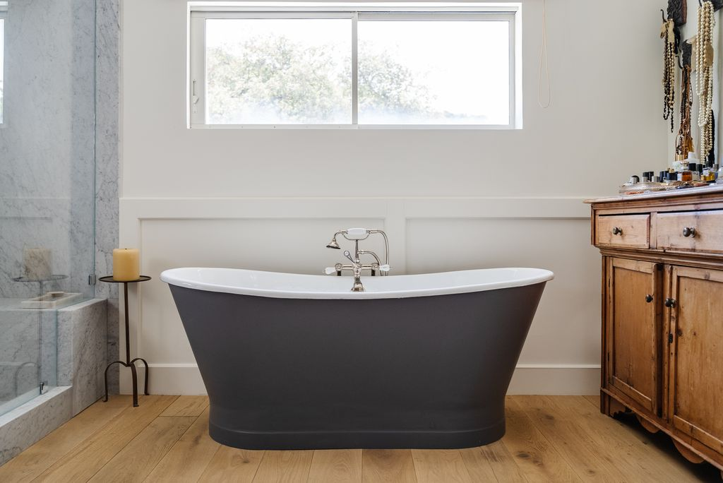 Across the room, a freestanding soaking tub sits beneath a horizontal picture window overlooking the trees outside.