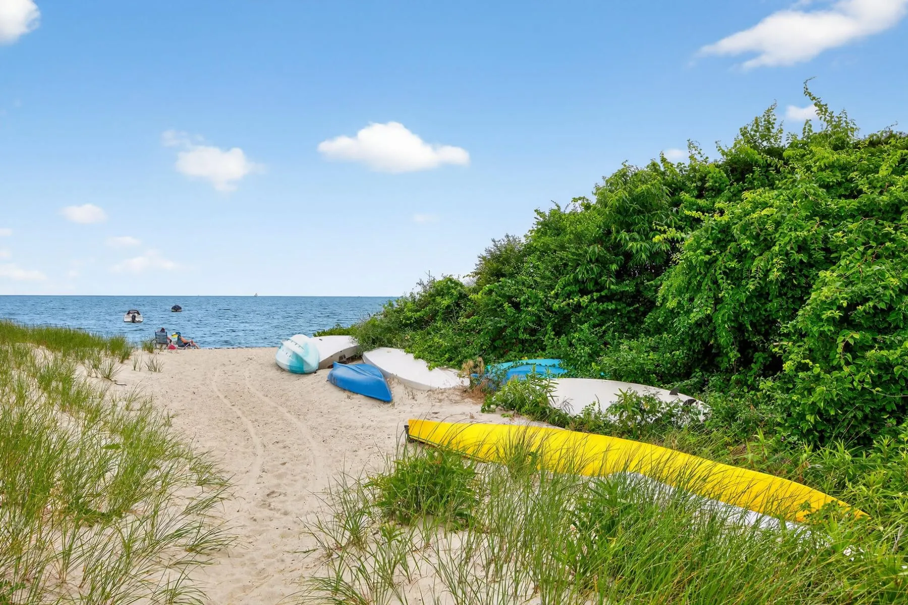 A sandy pathway winds through the dunes toward the water, where kayaks and paddleboards line the edge of the beach.
