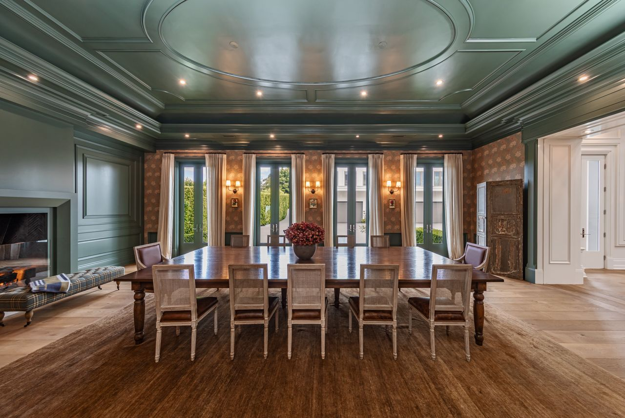 A long wood dining table sits in the center beneath recessed lighting, while tall French doors lined with soft drapery bring in natural light and open the room toward the grounds outside. 