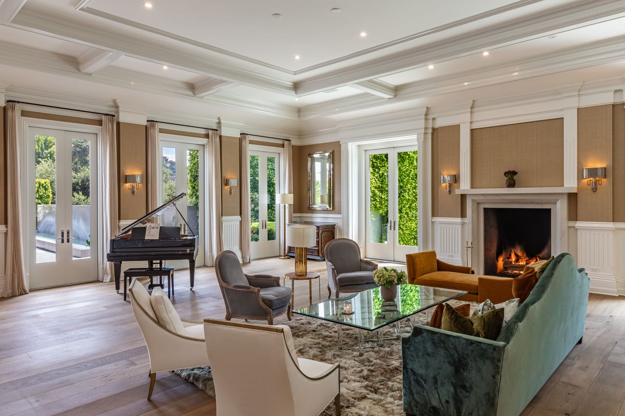 The seating arrangement centers around a large fireplace framed by crisp white millwork, with velvet sofas, armchairs, and a glass coffee table creating a balanced gathering space in the middle of the room.