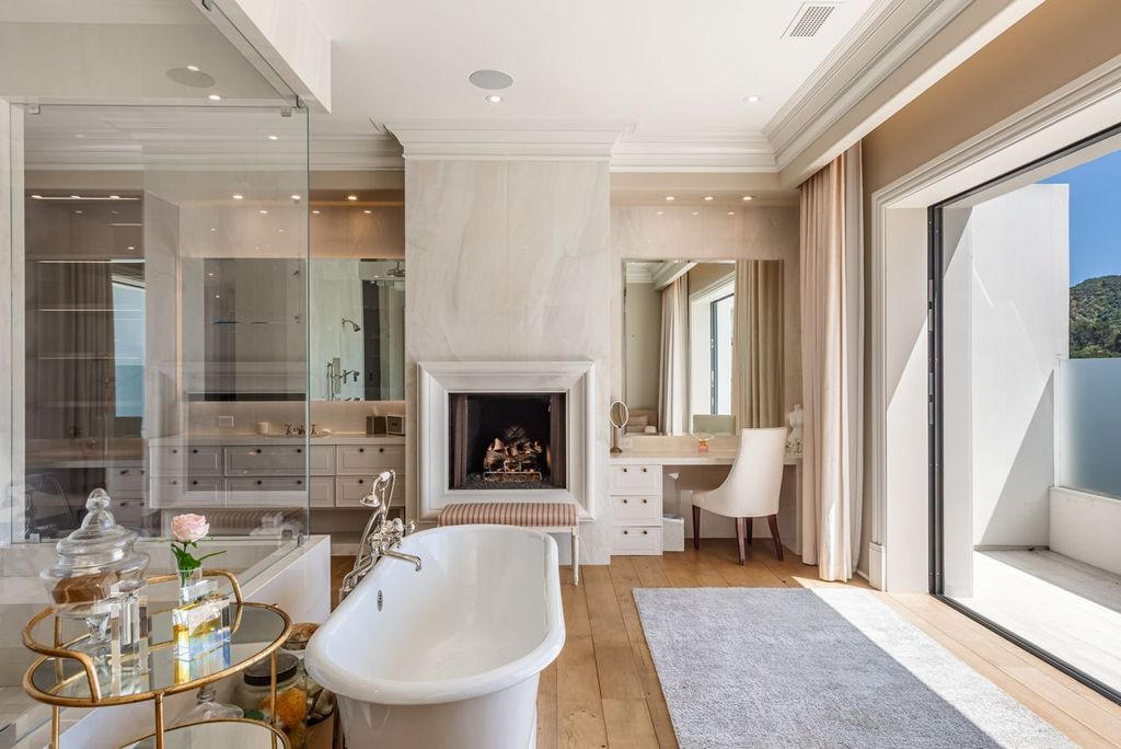 A freestanding soaking tub sits near the center of the room beside a fireplace and built-in vanity area, while floor-to-ceiling glass encloses an oversized walk-in shower with dual rainfall fixtures.