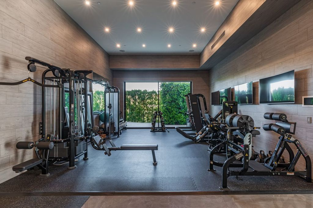 The gym takes on a more contemporary look with wood-paneled walls, rubber flooring, and full-height glass doors opening toward landscaped greenery outside. 