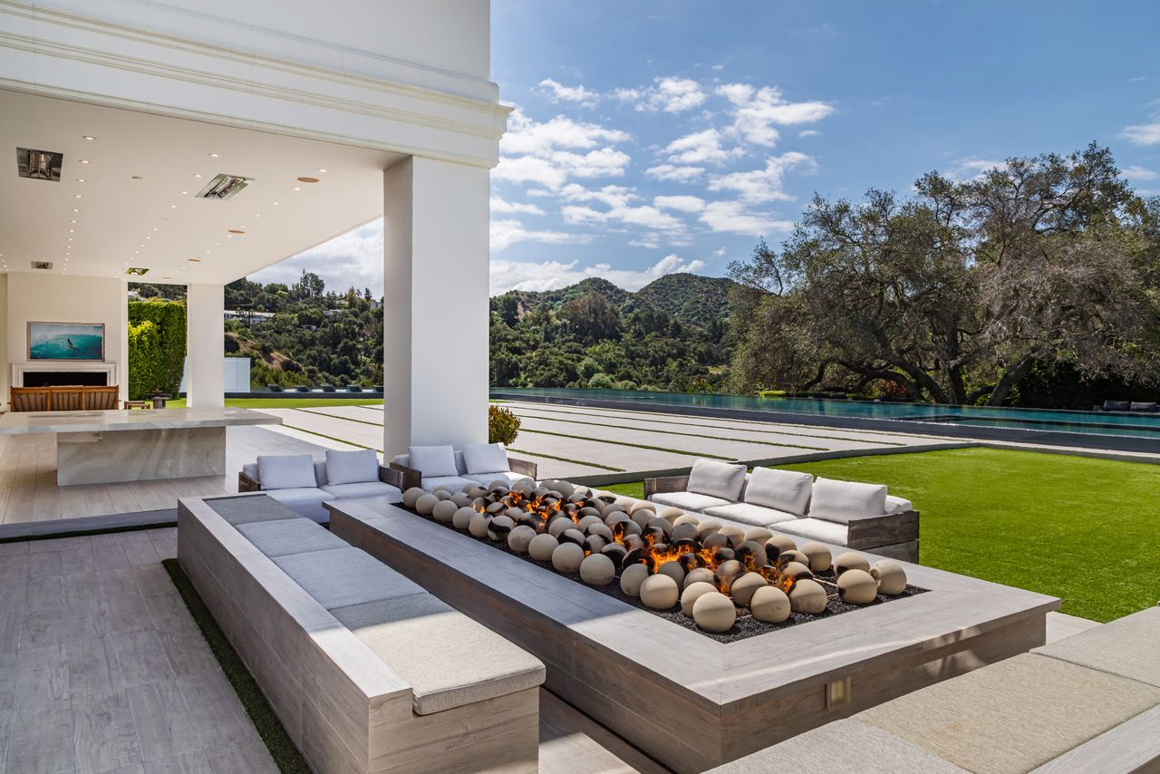 A few steps beyond the covered terrace, the backyard opens into a dedicated firepit lounge anchored by a long linear flame feature filled with smooth decorative stones. 