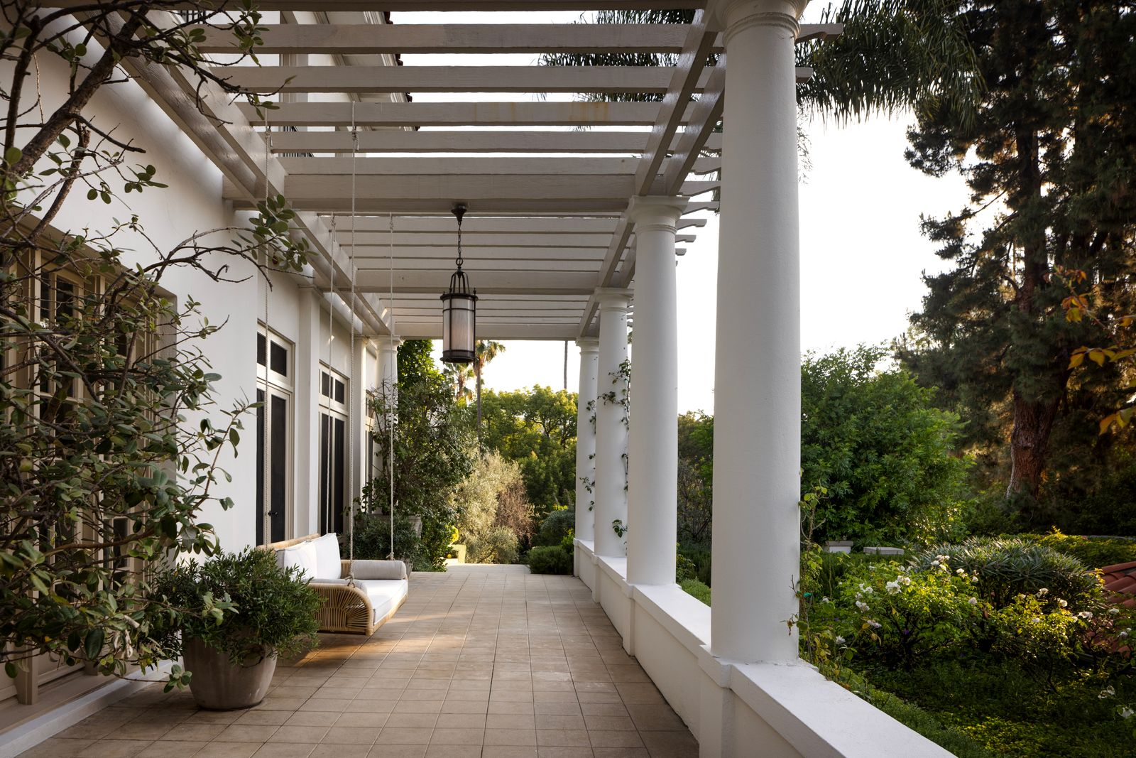 A column-lined veranda runs alongside part of the main residence, shaded by a pergola overhead and framed by climbing greenery and oversized lanterns. 