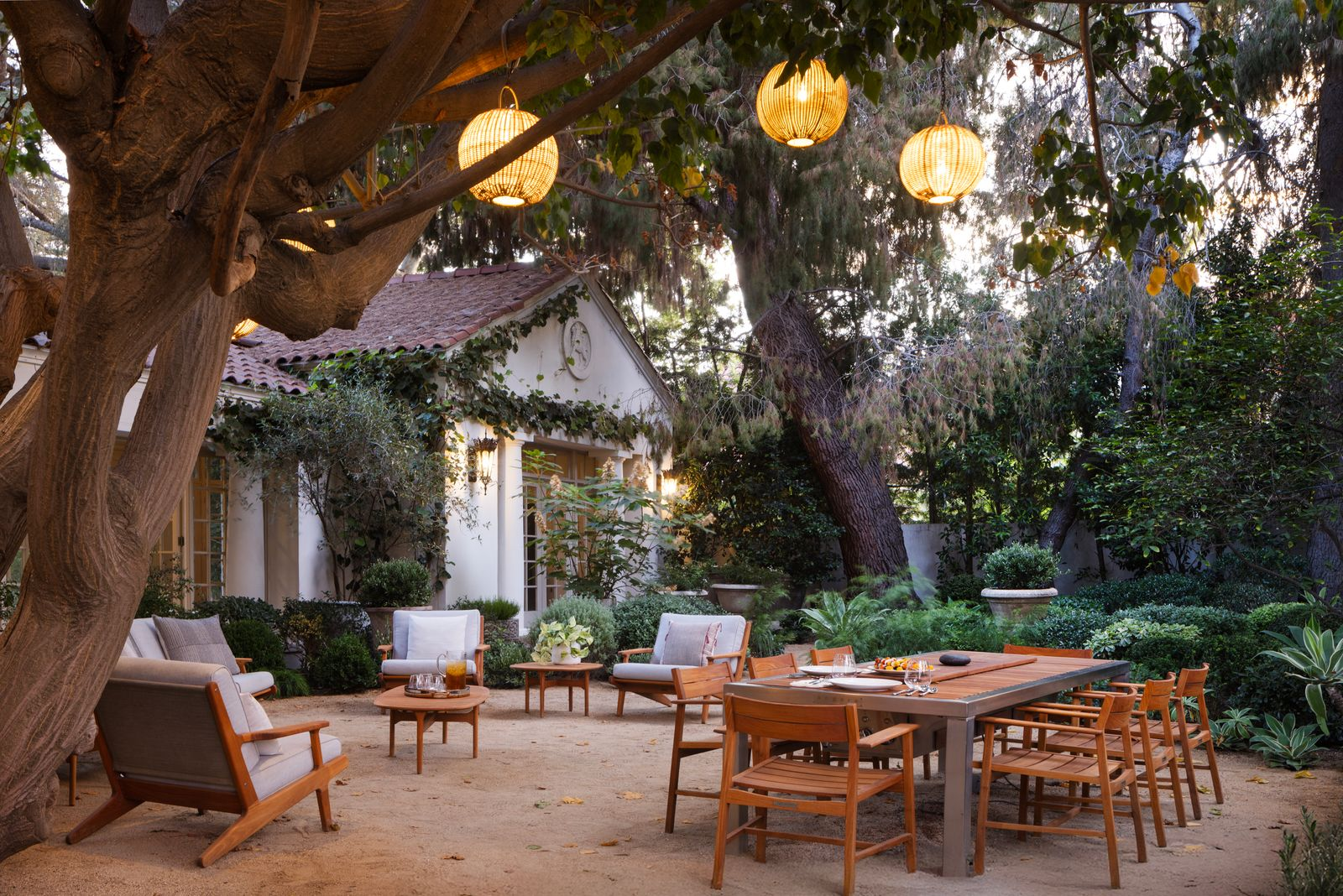 A long dining table is placed beside multiple lounge chairs arranged for conversation, creating separate zones for dining and casual gatherings beneath the trees.