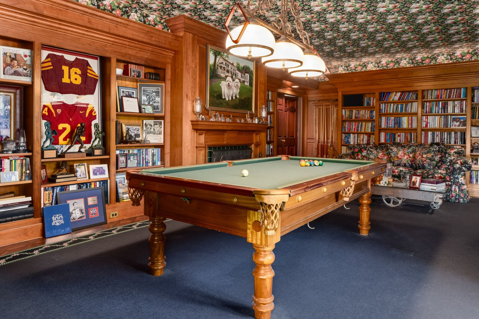 Rich wood paneling, built-in bookshelves, and a detailed patterned ceiling frame the space, while a classic billiards table sits in the center of the room beneath oversized pendant lights.