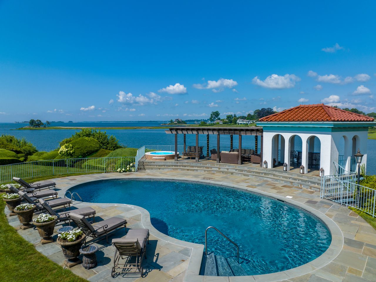 Positioned near the waterfront, the curved swimming pool is bordered by wide stone decking and rows of lounge chairs overlooking the bay. 