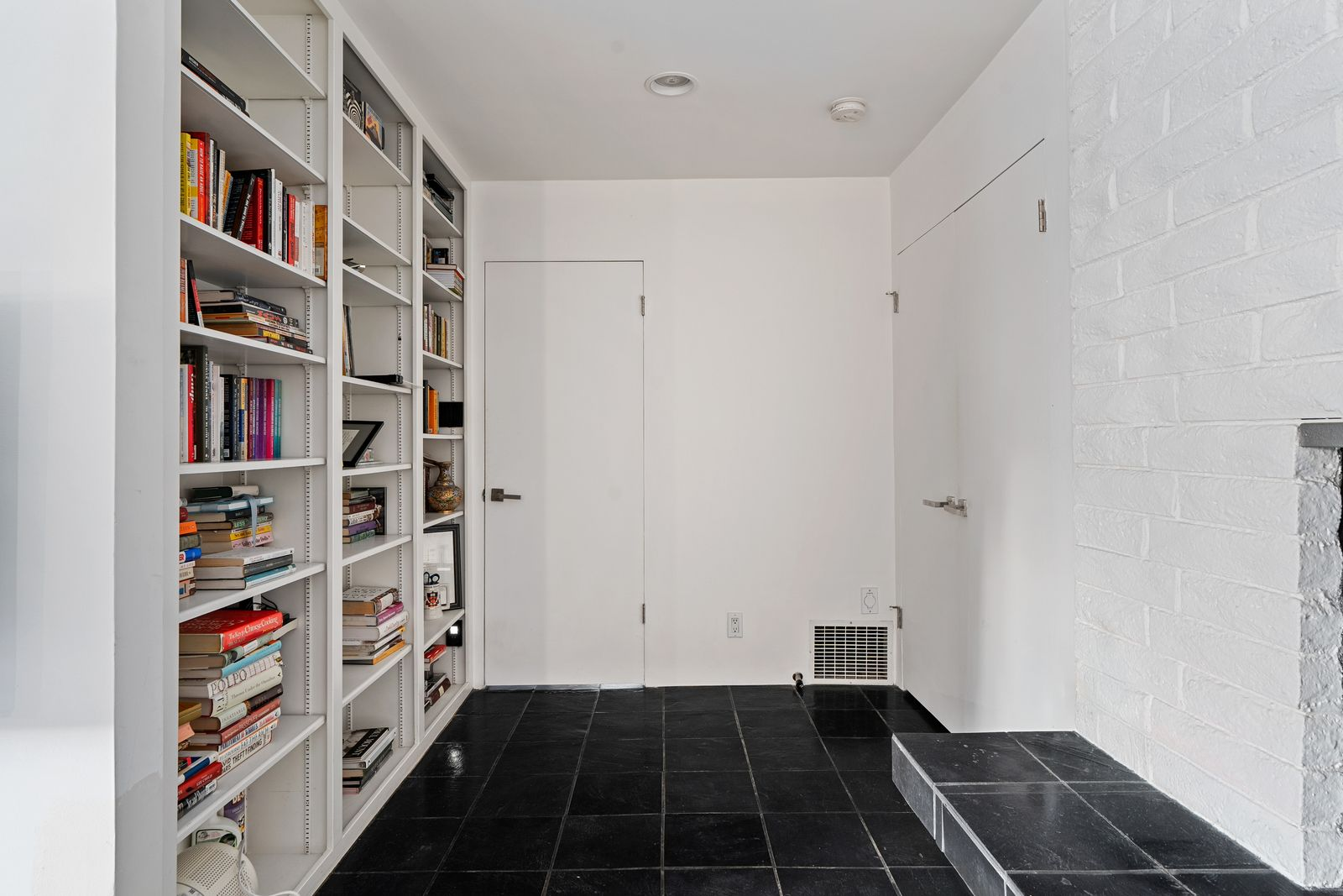 Just off the sleeping area, a compact library nook introduces another functional detail, with built-in shelving running floor-to-ceiling across from a white-painted brick fireplace wall.
