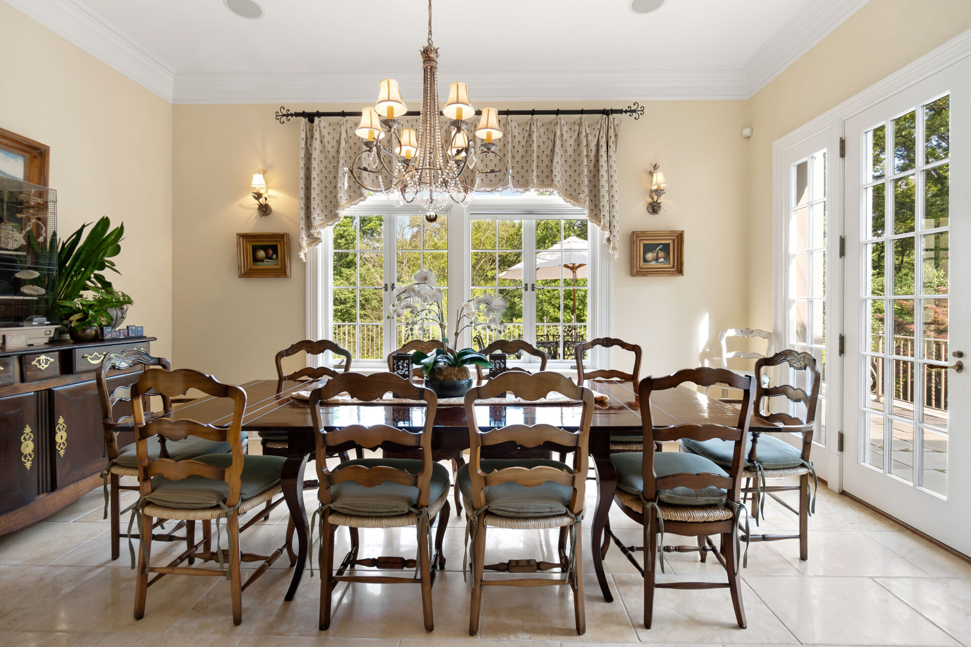 A large table sits beneath another chandelier, creating an everyday dining area that overlooks the grounds and provides direct access to an outdoor terrace.