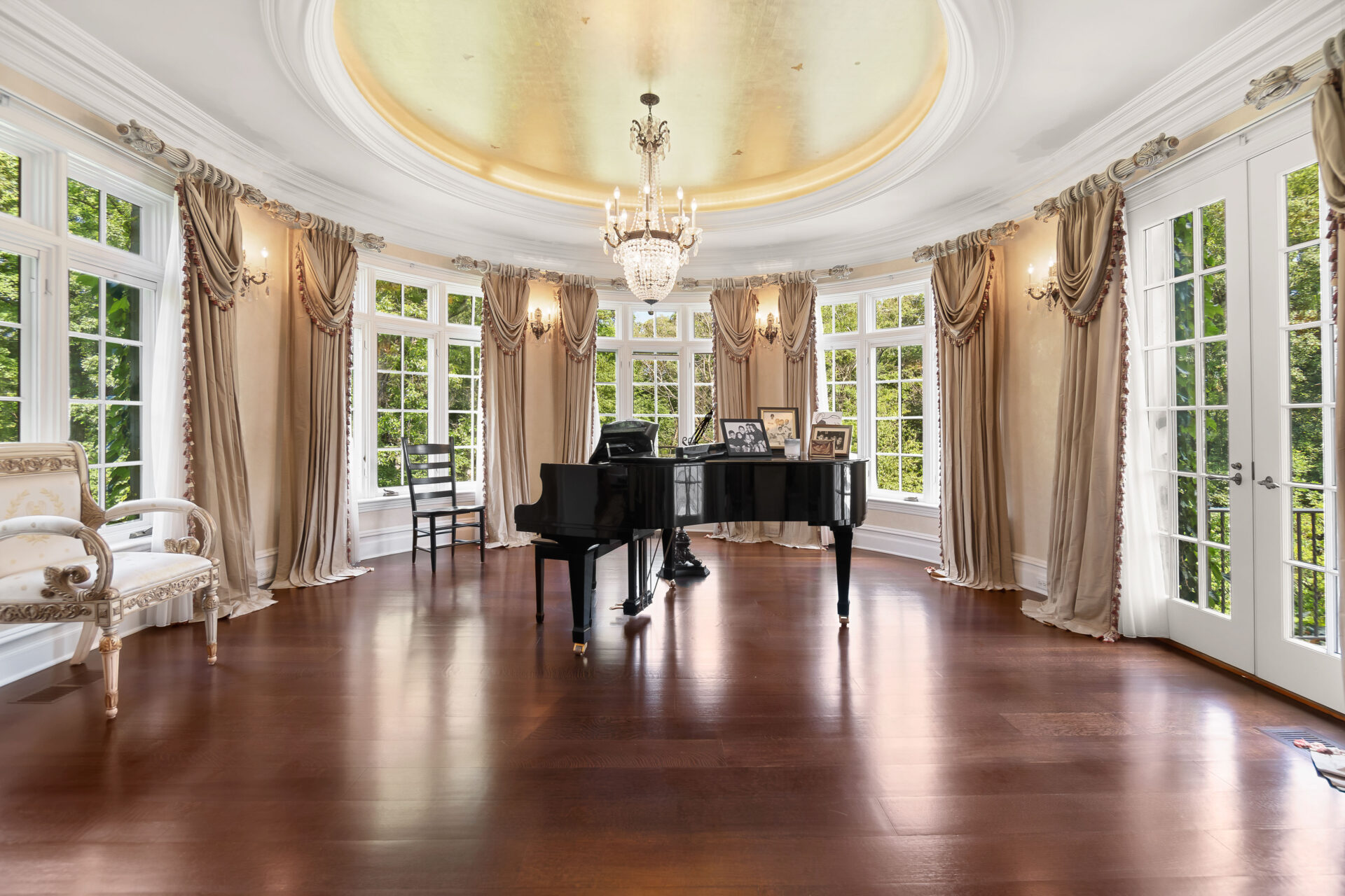 A grand piano occupies the middle of the space, while a ring of tall windows and French doors surrounds the room with views of the property.