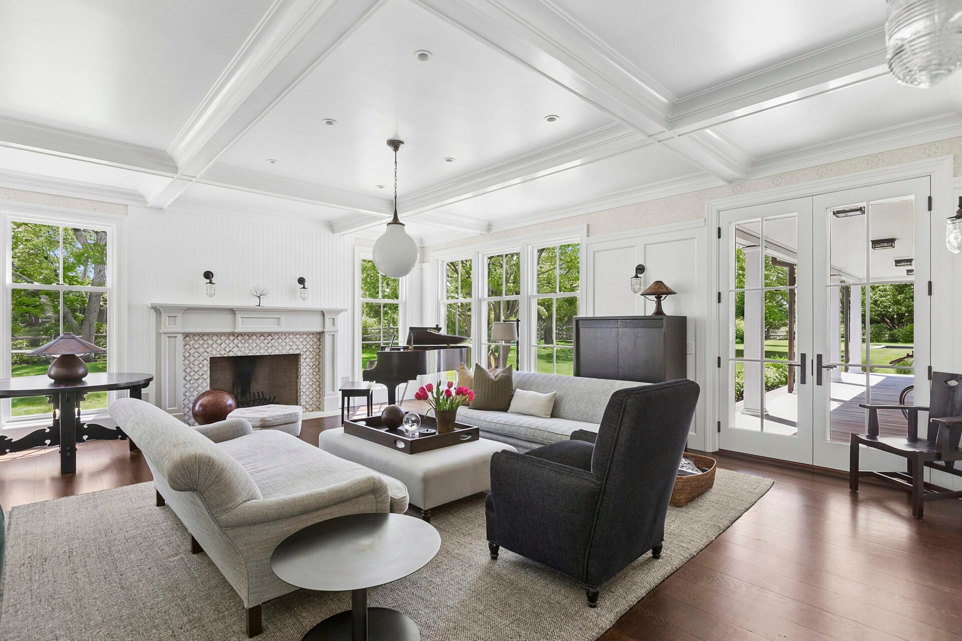 At the center of the space, curved upholstered sofas and a large ottoman anchor the room across from a tile fireplace and grand piano.