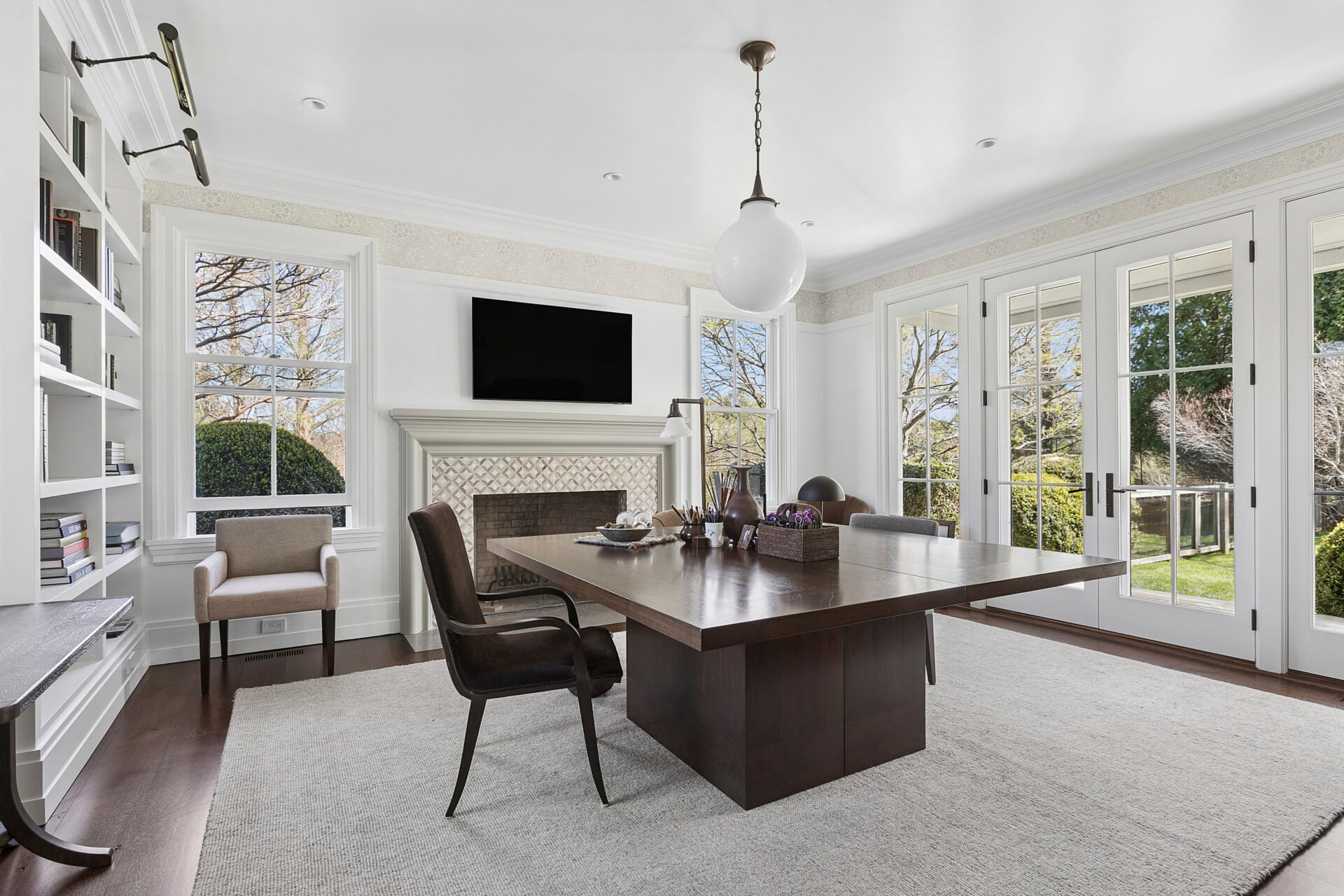 The second space functions as a library or home office, with a wood desk positioned in front of a fireplace, built-in shelving, and French doors opening directly to the outdoors.
