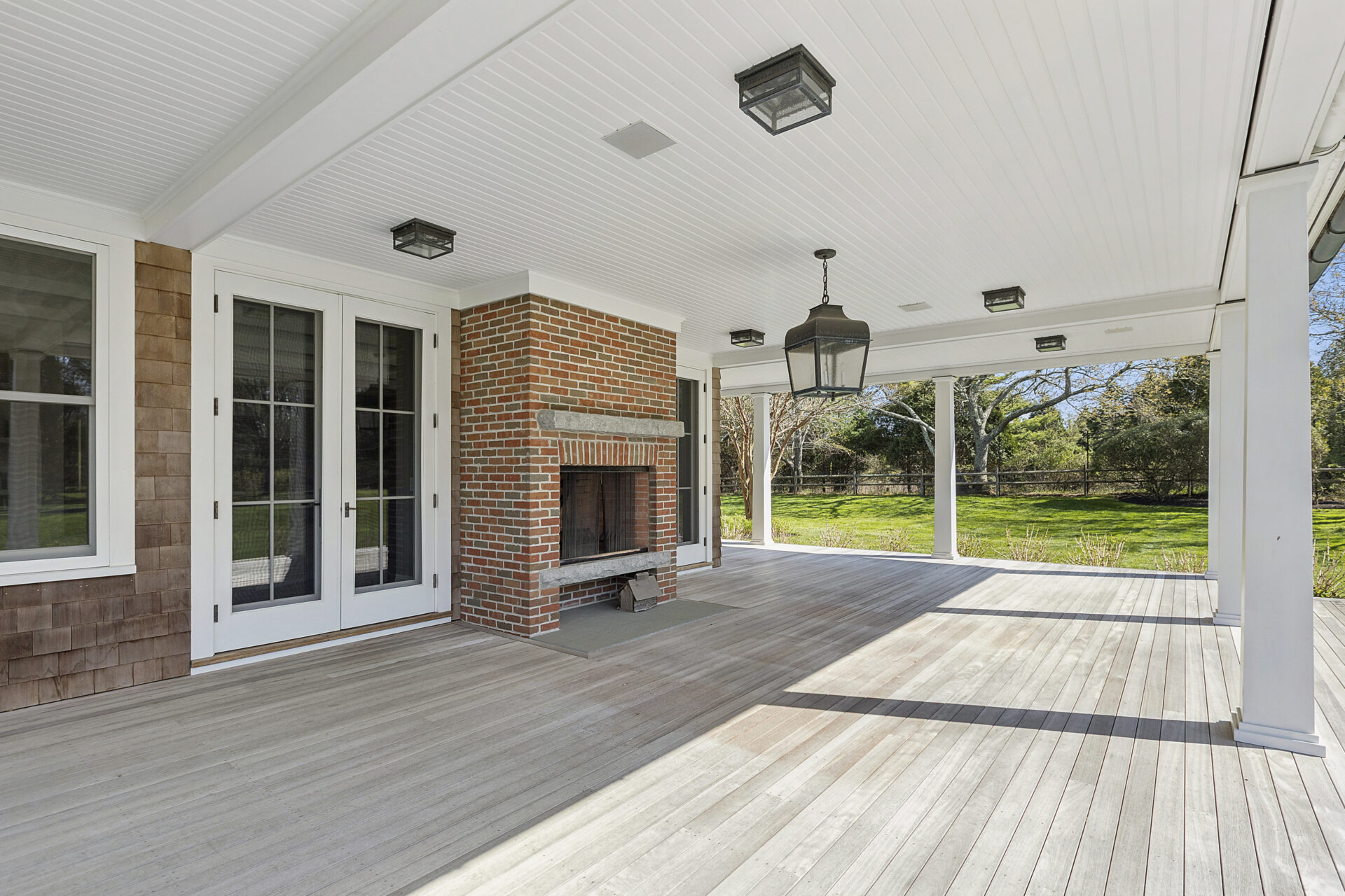 White columns support a beadboard ceiling fitted with lantern-style lighting, while a brick fireplace creates the perfect lounge spot along one wall.