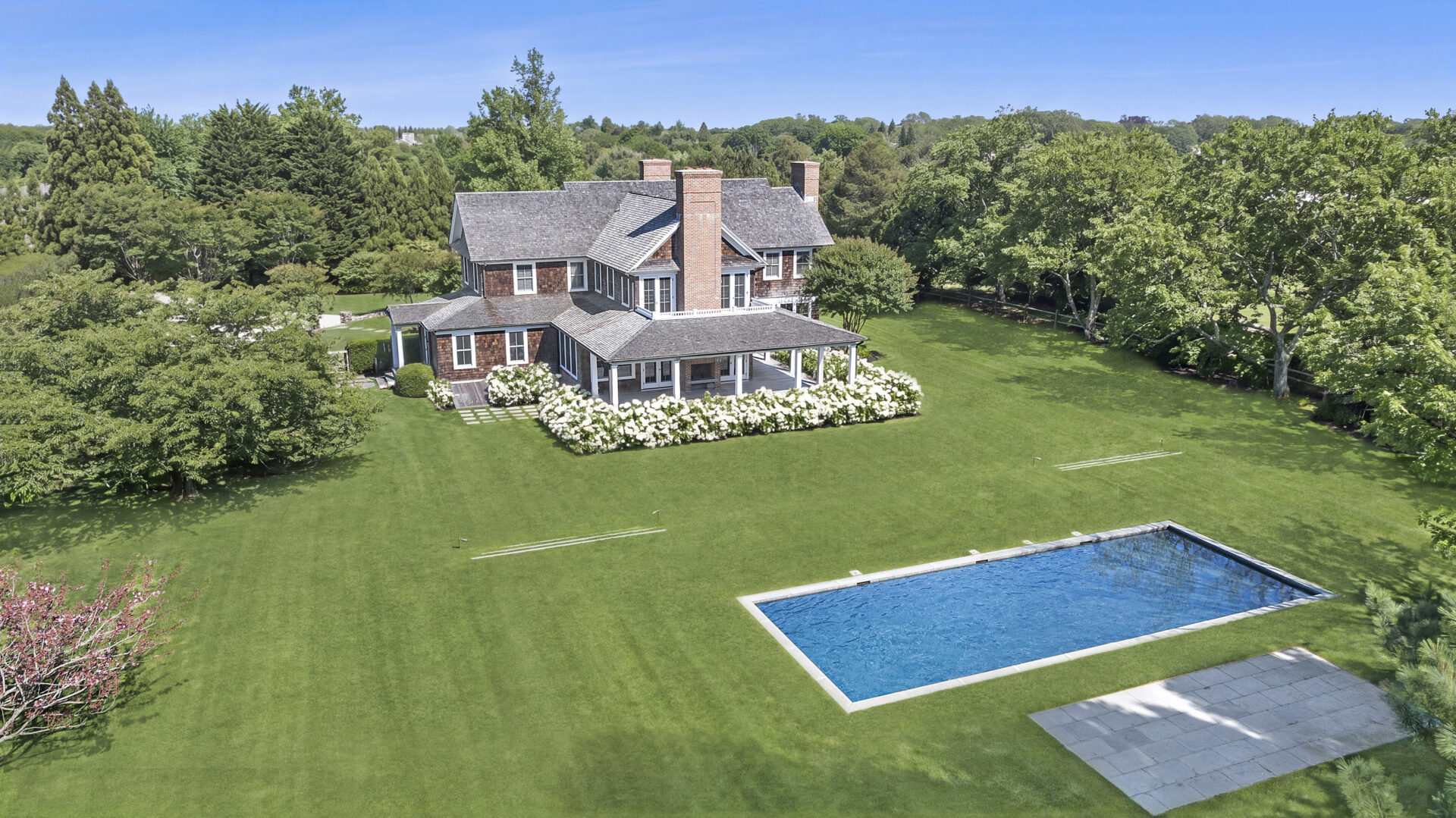A border of mature specimen trees, flowering hydrangeas, and curated plantings frames the pool area.