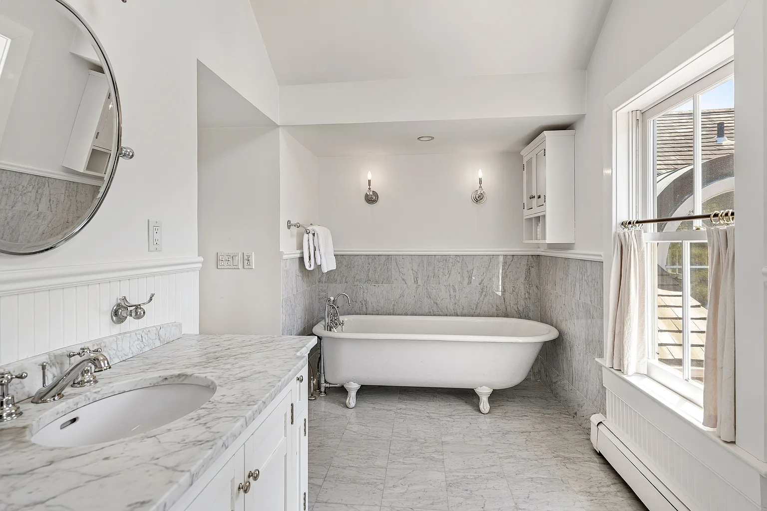 A marble-topped vanity sits opposite a freestanding clawfoot soaking tub positioned beneath a large window.