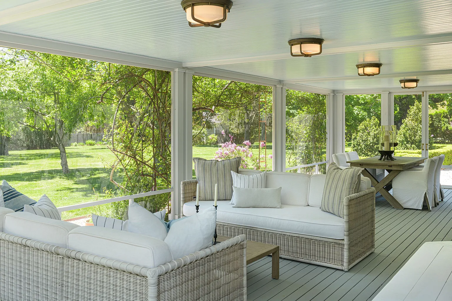 At one end, a lounge setting with woven sofas and cushioned seating creates a comfortable place for conversation, while a large dining table is placed at the opposite side of the porch.