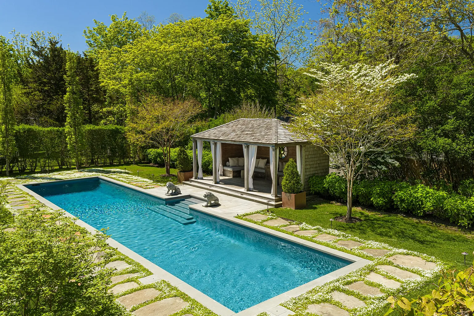 Positioned alongside the pool is a covered cabana designed as an outdoor living room open on multiple sides and finished with draped curtains and built-in seating.