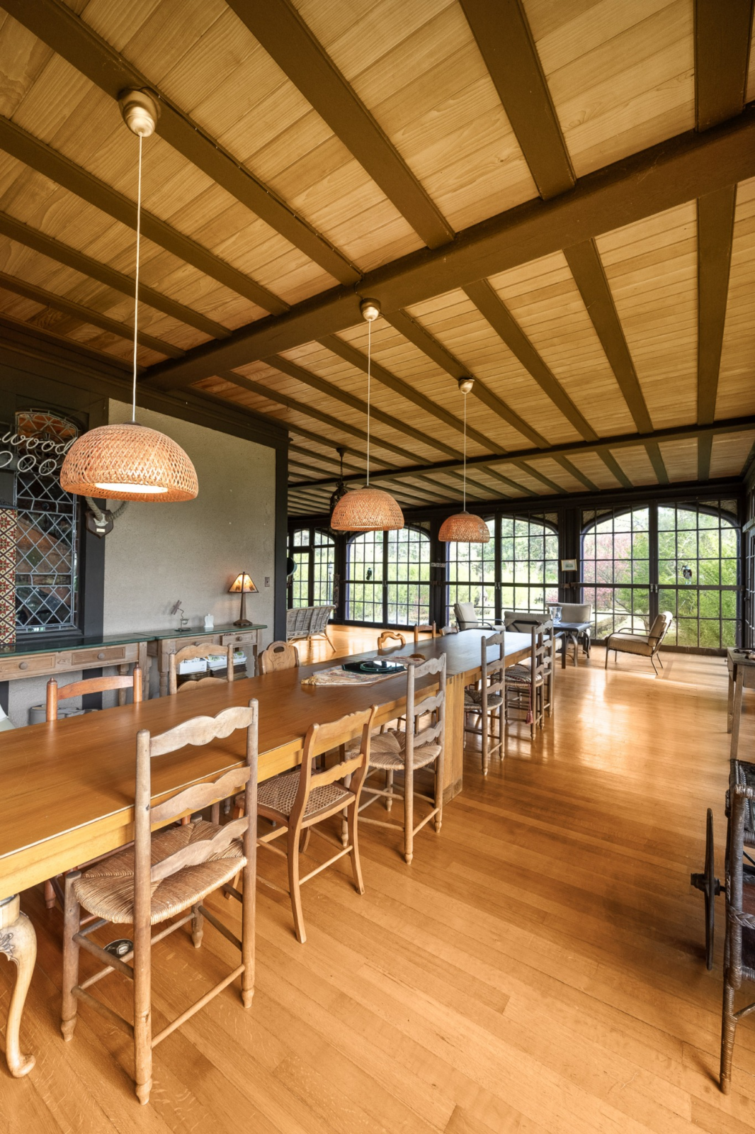 Positioned between indoor and outdoor living spaces, the room features a long farmhouse-style table beneath exposed ceiling beams and woven pendant lights.