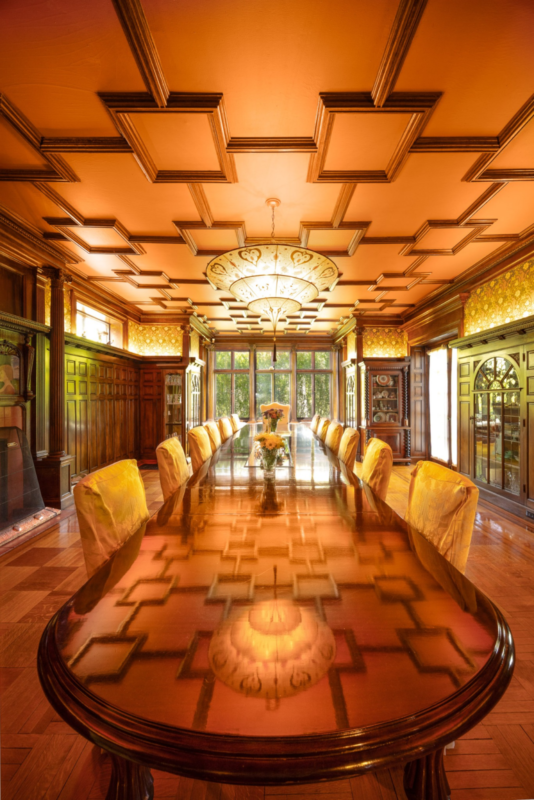 Paneled wood walls, built-in cabinetry, decorative columns, and an intricate geometric ceiling frame a polished dining table sized for large gatherings. 