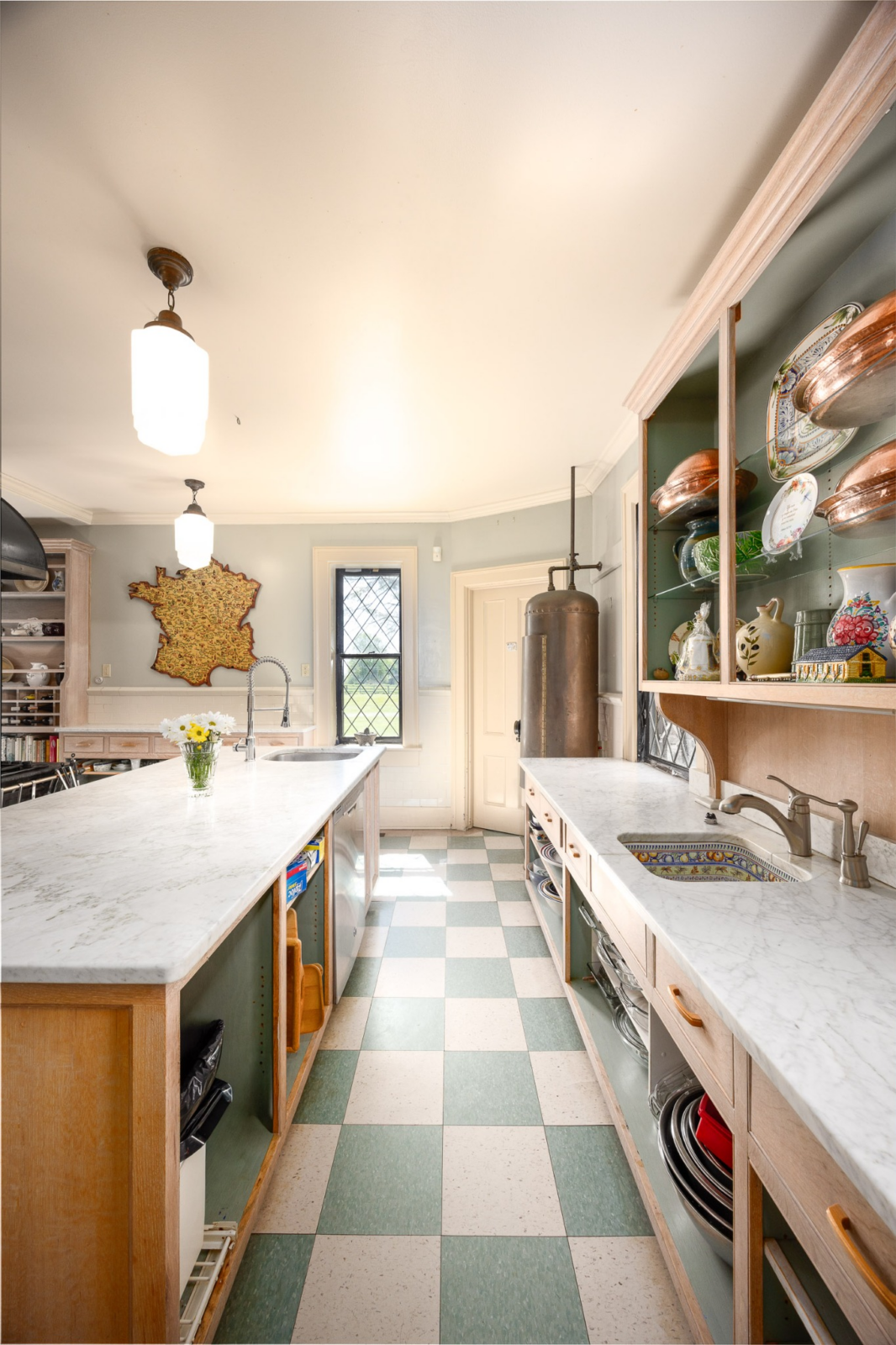 Open shelving, built-in storage, and display cabinets showcase copper cookware and collected pieces, while the checkerboard flooring and oversized farmhouse-style sink add to the kitchen’s distinctive personality.