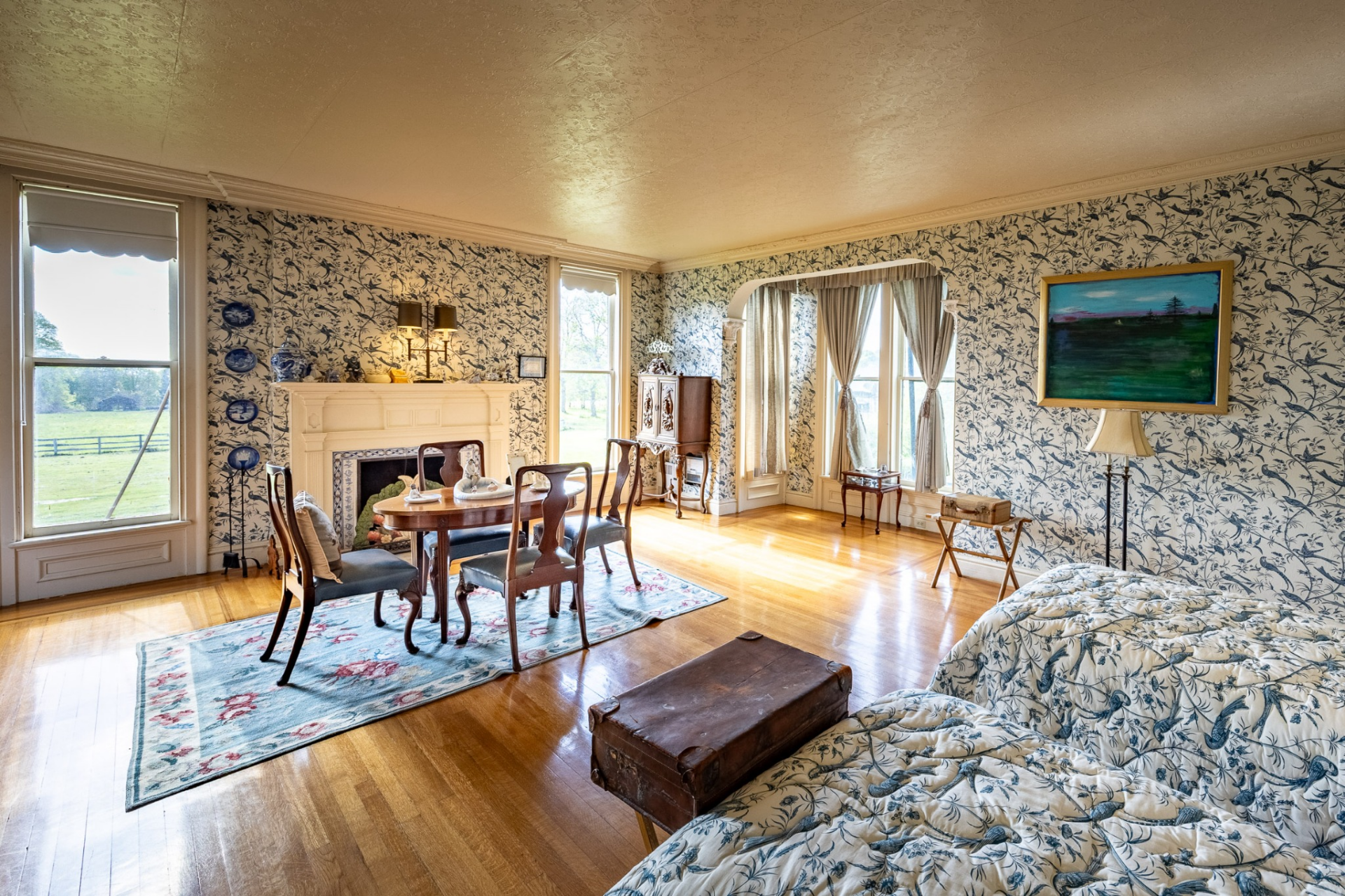 A decorative fireplace is framed by period millwork within the same line as a dining table for four, and two twin beds with blankets that match the wallpaper.