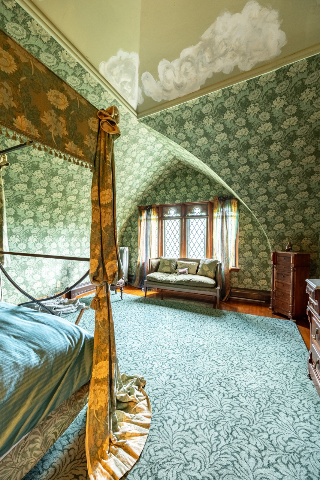 A four-poster canopy sits across from window seating beneath the arched dormer, while carved wood trim, leaded-glass windows, and decorative plaster ceiling motifs reinforce its early 20th-century craftsmanship.