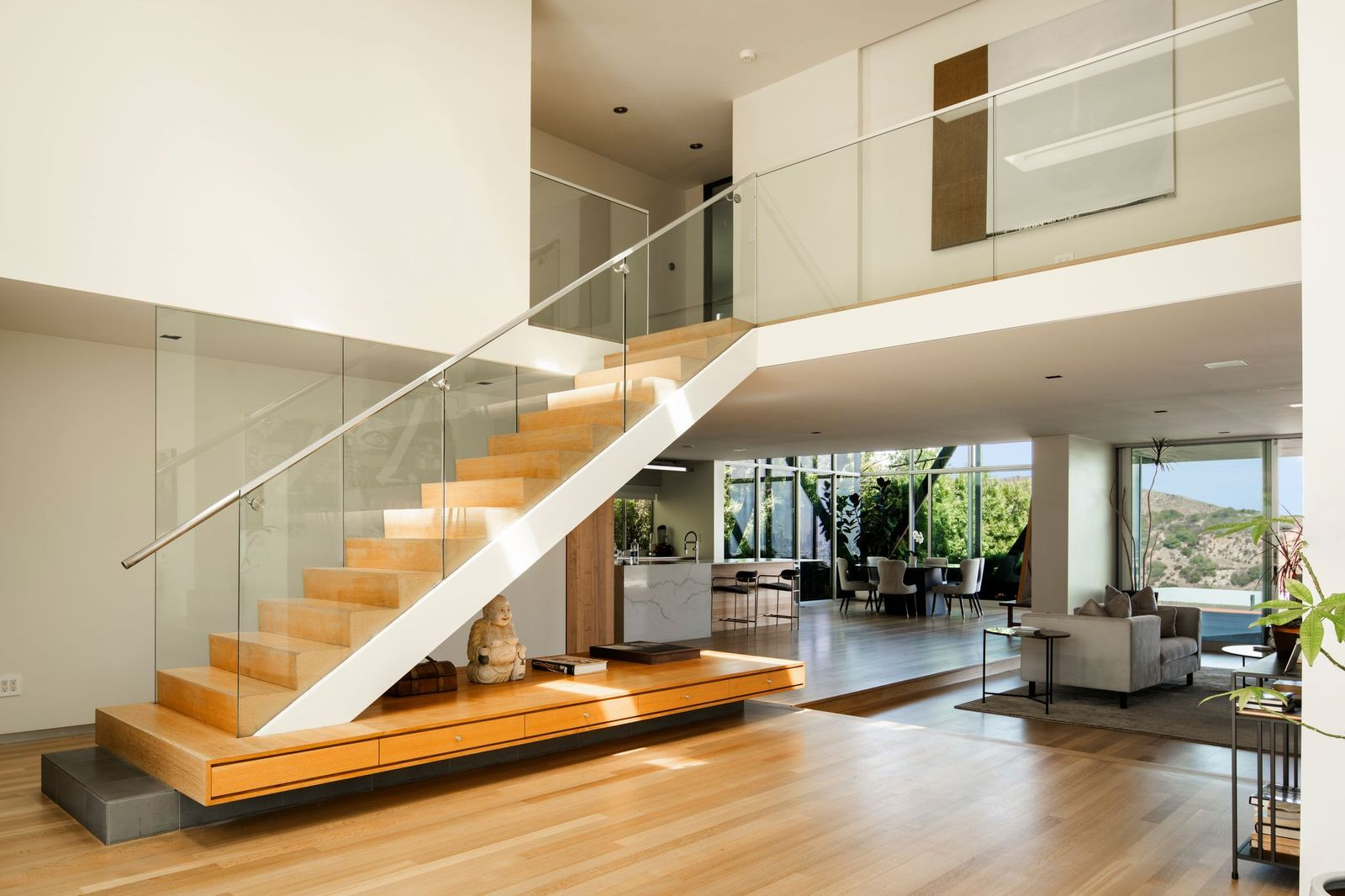 To the right, a floating staircase lined with a glass railing becomes the focal point of the room, as it stretches over a built-in wood bench, adding both functionality and warmth.